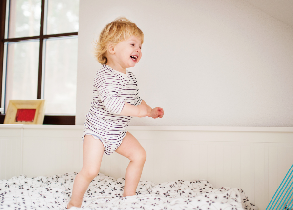Child with blonde hair wearing a stripey romper smiles while standing on a mattress