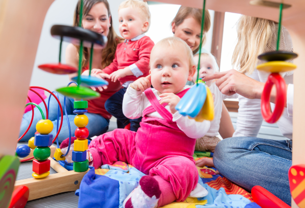 Baby wearing bright pink dungarees points to ear while surrounded by wooden toys