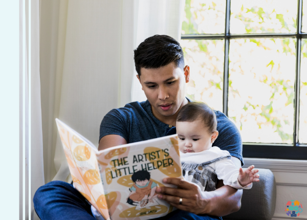 An adult reading to a baby, the book is "The Artist's Little Helper"