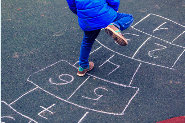 Child in blue coat and jeans playing hopscotch that has been drawn on the floor with chalk. Child is mid leap with his right foot in the air. 