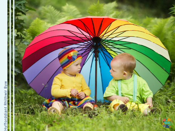 Two babies sitting under a rainbow umbrella