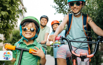 Group of people on bikes, at the front is a young child smiling while holding a scooter