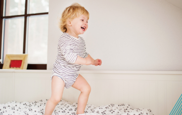 Child with blonde hair wearing a stripey romper smiles while standing on a mattress