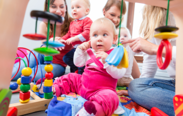 Baby wearing bright pink dungarees points to ear while surrounded by wooden toys