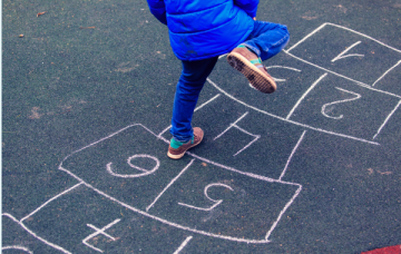 Child in blue coat and jeans playing hopscotch that has been drawn on the floor with chalk. Child is mid leap with his right foot in the air. 